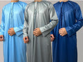 Three men wearing traditional Emirati thobes in blue, gray, and black colors, featuring a shiny fabric and a tassel design.